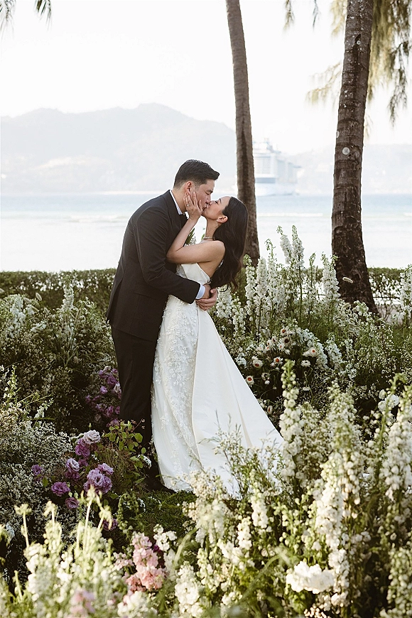 Wedding kiss portrait of bride and groom kissing, bride in strapless lace dress with pearl necklace amid garden flowers, ocean and palm trees behind