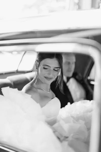 Bride portrait in car with a strapless wedding dress, looking down with a soft smile as the groom sits blurred in the background