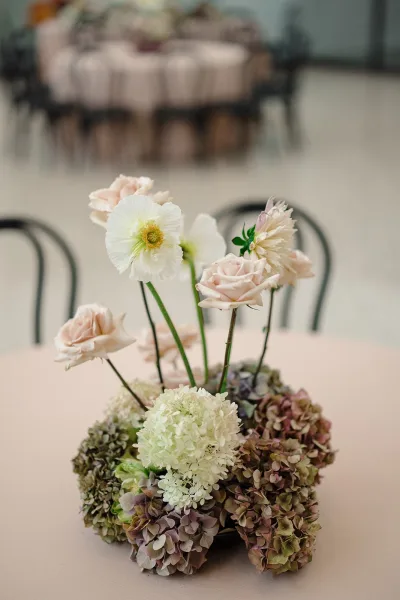 Wedding centerpiece with hydrangea wedding centerpiece blooms, blush roses and a white poppy in bud vases on a round tablecloth in reception room