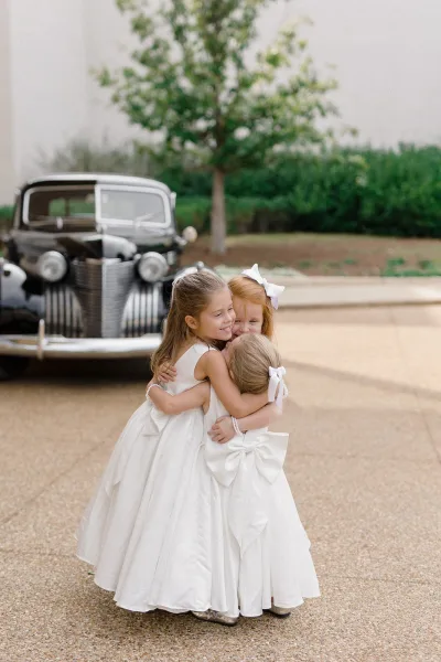 Flower girl portrait of three girls hugging in white dresses with hair bows and pearl bracelets beside a vintage car on a gravel driveway