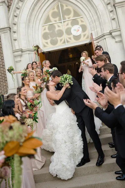 Wedding kiss as the bride and groom dip on church steps, bouquet in hand, while bridesmaids and groomsmen cheer at the doorway