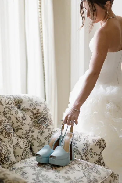 Bride getting ready in a strapless tulle gown, fastening blue platform heels beside a vintage floral armchair in soft window light