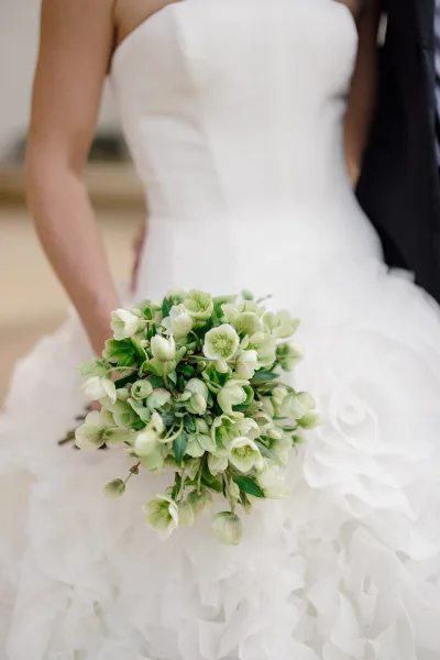 Bridal bouquet of green and white bouquet flowers held by bride in strapless tulle gown beside groom in black suit, neutral blur backdrop