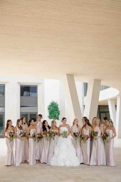 Bridesmaids portrait with bride in blush satin dresses holding green-and-white bouquets, posed by modern white columns and windows outdoors