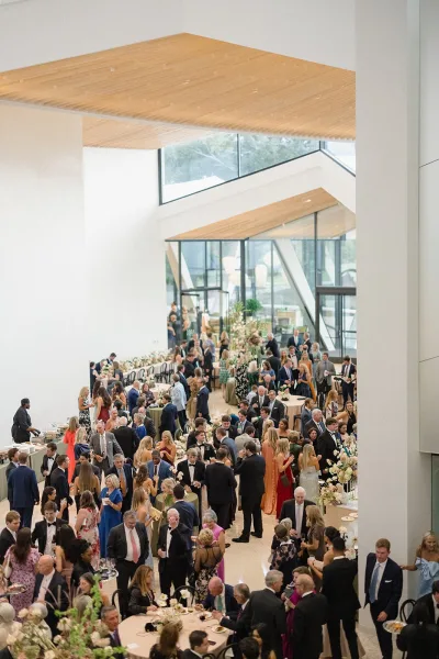 Wedding reception cocktail hour with black-tie guests mingling around round tables, floral centerpieces, in a bright modern venue with tall windows