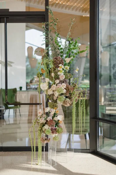 Wedding floral arrangement with roses, anthurium, and hydrangea on clear acrylic pedestals by glass doors in a string-lit reception space