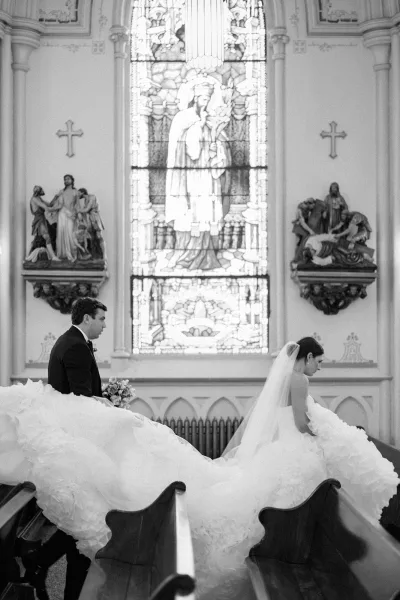 Ceremony moment as bride in a cathedral veil and strapless ball gown sits in church pews, groom in tux with bouquet near stained glass window