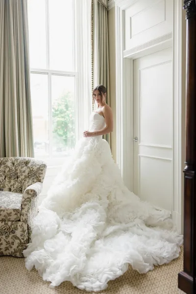 Bridal portrait of a bride in a strapless wedding dress with ruffled tulle skirt and long train, standing by a tall window in a hotel suite