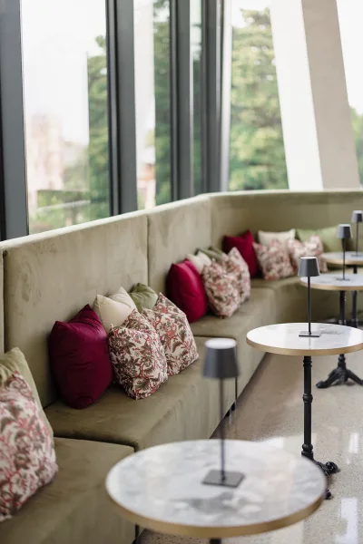 Lounge seating area with built-in banquette, velvet pillows, marble bistro tables and lamps by floor-to-ceiling windows with greenery outside