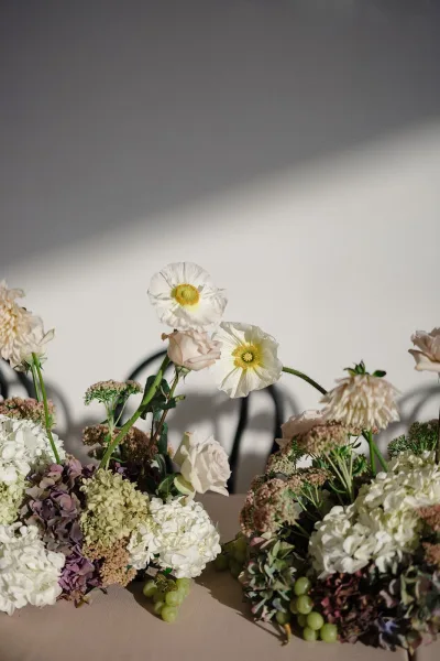 Wedding floral centerpiece with hydrangea centerpiece blooms, poppies, blush roses, greenery, and green grapes on a sunlit table by a white wall