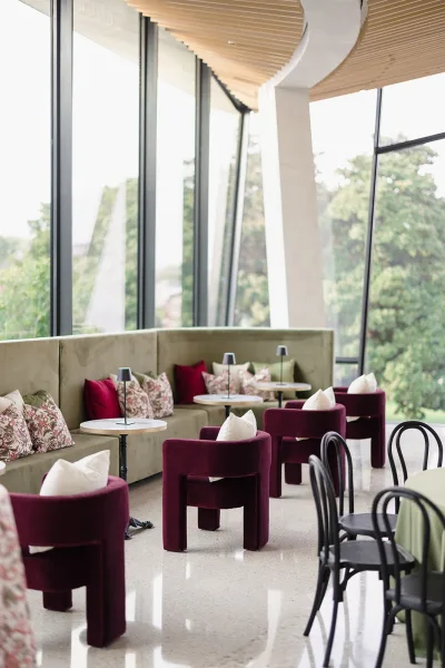 Modern lounge seating with burgundy chairs and a banquette sofa, styled with pillows and lamps, set by floor-to-ceiling windows and trees