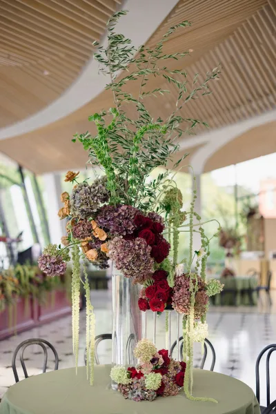Wedding centerpiece in a tall clear vase with hydrangea and red roses, cascading greenery on a green tablecloth in a bright reception room