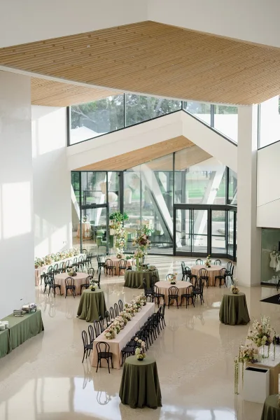 Reception layout with round tables and a long banquet table, floral centerpieces and greenery garland in a modern white venue with windows