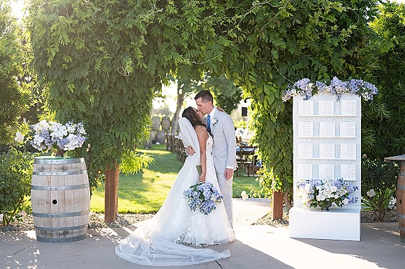 Wedding kiss portrait of bride and groom kissing under a greenery arch, her long veil draped behind, holding a blue-and-white bouquet outdoors