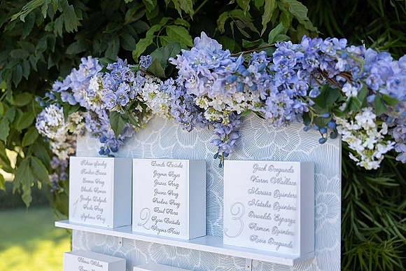 Wedding seating chart with escort cards display on a white shelf, topped with blue and white flowers and greenery against garden lawn backdrop