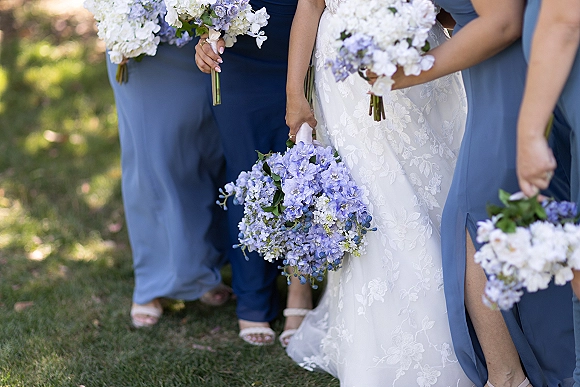 Bridal party portrait of bride with bridesmaids in dusty blue dresses holding blue and white bouquets on a green lawn outdoors