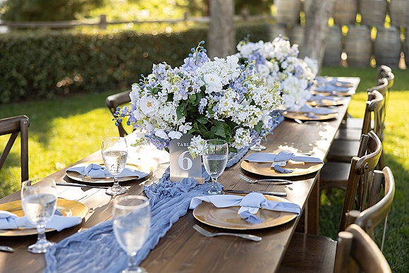 Reception tablescape with an outdoor reception table on a farmhouse wooden table, blue runner, gold chargers, florals, and barrel-lined lawn backdrop