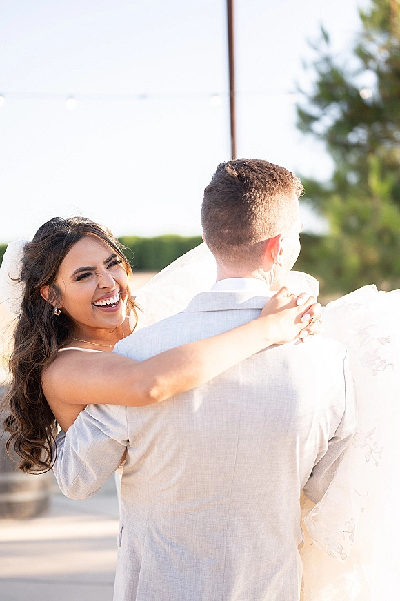 Wedding couple portrait with groom holding a laughing bride in a veil and strapless dress, outdoors among trees under bright sky