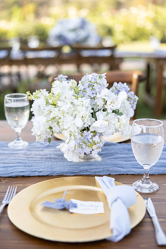Reception tablescape with a blue table runner, white and light blue floral centerpiece, and gold charger plates on a wooden table outdoors