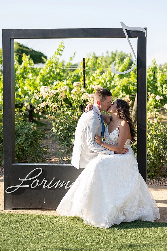 Wedding kiss portrait of bride and groom kissing in a giant acrylic frame with calligraphy, set on a sunny vineyard path, blue sky behind