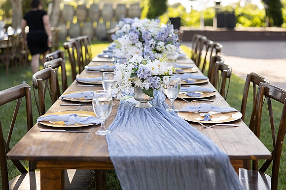 Reception tablescape with outdoor wedding table setting on a wood farm table, blue runner and compote florals on a sunny lawn patio