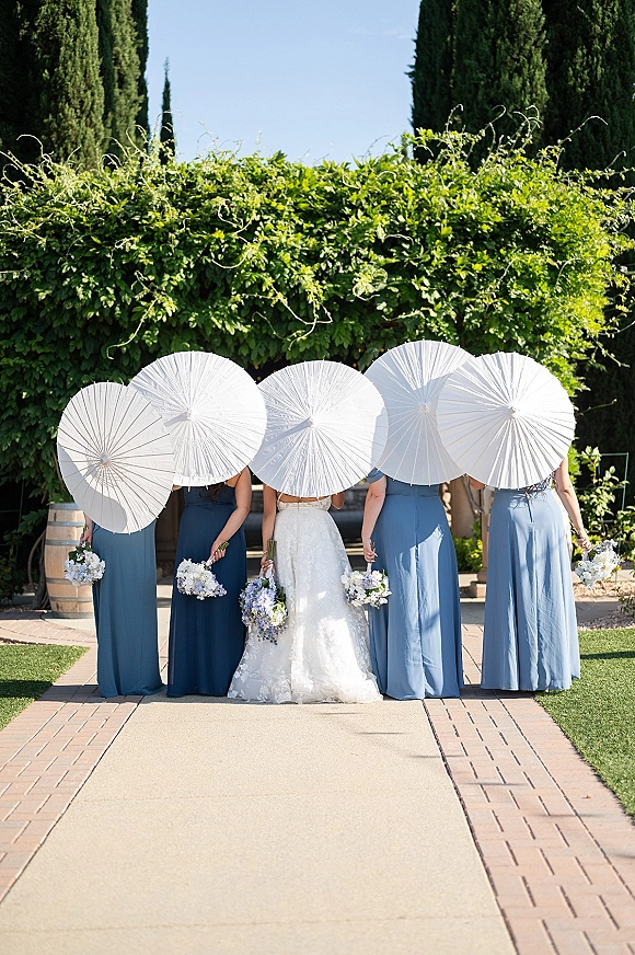 Bride with bridesmaids holding bouquets, shaded by white paper parasols, walking along a garden path beneath tall trees and blue sky
