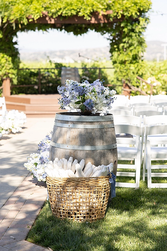 Ceremony aisle decor with a wine barrel floral arrangement of blue and white blooms beside a program basket on an outdoor lawn aisle