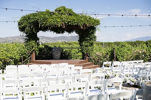 Ceremony setup for an outdoor wedding ceremony with greenery arch and string lights, white chairs, and floral aisle markers in a vineyard with mountains