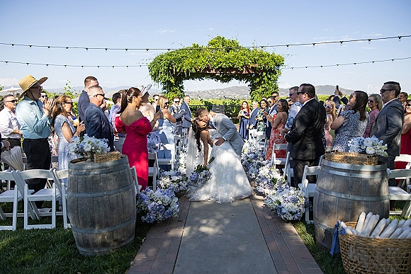 Ceremony kiss as bride and groom embrace beneath a greenery wedding arch with string lights, guests cheering along a flower-lined aisle on an outdoor lawn