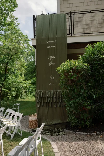 Wedding welcome sign as fabric wedding signage with ruffle trim, greeting guests along a gravel garden path by trees and a house balcony.