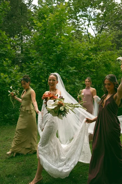 Bride and bridesmaids in a walking bridal party, sharing a candid moment as the bride’s veil trails across a grass lawn beside forest trees.