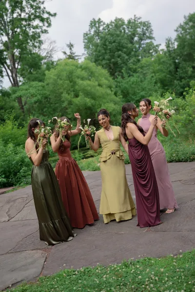Bridesmaids portrait in mismatched bridesmaid dresses, each holding small calla lily bouquets along a stone path amid garden greenery.