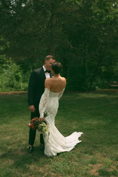 Couple portrait of a bride and groom embracing in an outdoor couple portrait on a grass lawn, the bride in a lace wedding dress by woodland trees.