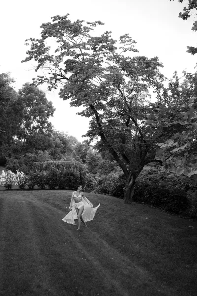 Bridal boudoir portrait of a bride in lingerie, wrapped in a light bridal robe, standing barefoot on a grass lawn edged by trees.