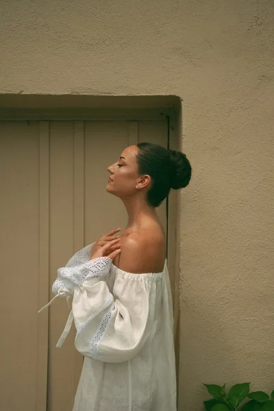 Bridal portrait of a bride in side profile wearing an off-the-shoulder dress, posed by a stucco wall with a paneled door and green leaves.