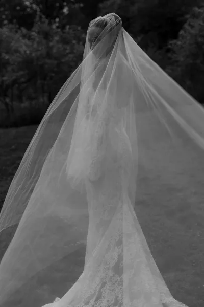 Bridal portrait of a bride under veil, her lace wedding dress visible, framed by garden trees and a neat grass lawn.