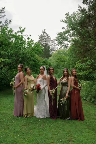 Bridal party portrait of a bride with bridesmaids, her classic veil framed by garden trees and lush greenery under an overcast sky.