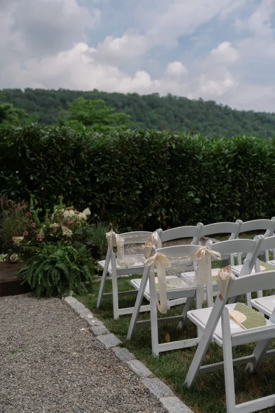 Ceremony seating featuring reserved chair signs, arranged in a garden setting with a hedge backdrop, distant hills, and a cloudy sky.