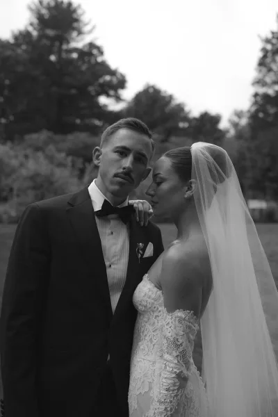 Couple portrait showing a bride and groom pose, her lace sleeves framed by garden trees on a lawn under an overcast sky.
