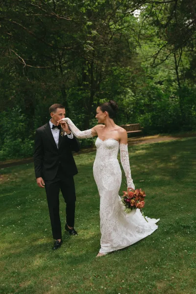 Couple portrait of a bride and groom walking together, the bride holding a bouquet, across a grass lawn by garden trees and a wooden bench.