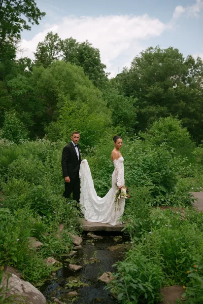 Couple portrait of a bride holding bouquet in a lace wedding dress beside her groom, set in garden greenery by a creek and footbridge.