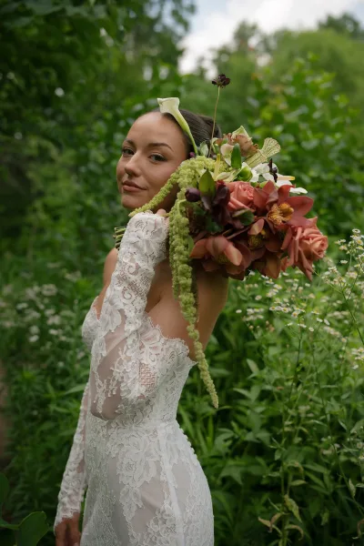 Bridal portrait of a bride with bouquet in a lace wedding dress, standing amid garden greenery and wildflowers beneath open sky.