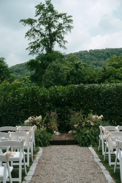 Outdoor ceremony setup with a garden ceremony aisle lined with white chairs along a gravel path, framed by hedges and a distant hillside.
