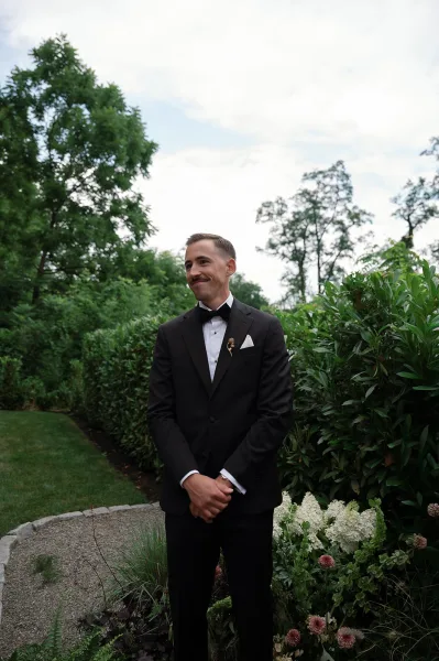 Groom portrait of a black tux groom adjusting his bow tie on a gravel path framed by garden hedges and trees.