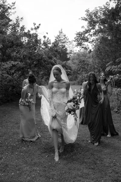 Bride and bridesmaids in a bridal party walking across a garden lawn, the bride cradling a classic bridal bouquet beneath leafy trees.