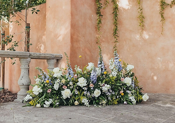 Wedding floral arrangement with white roses and blue delphinium, blush blooms and greenery on a stone patio by a stucco wall