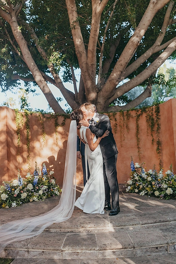 Wedding kiss portrait of bride and groom kissing, her long veil trailing beside floral ground arrangements in a garden stone patio setting
