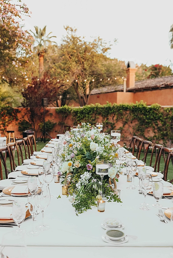 Reception tablescape on a long banquet table wedding setup with floral garland, floating candles, gold chargers, and string lights on a garden lawn