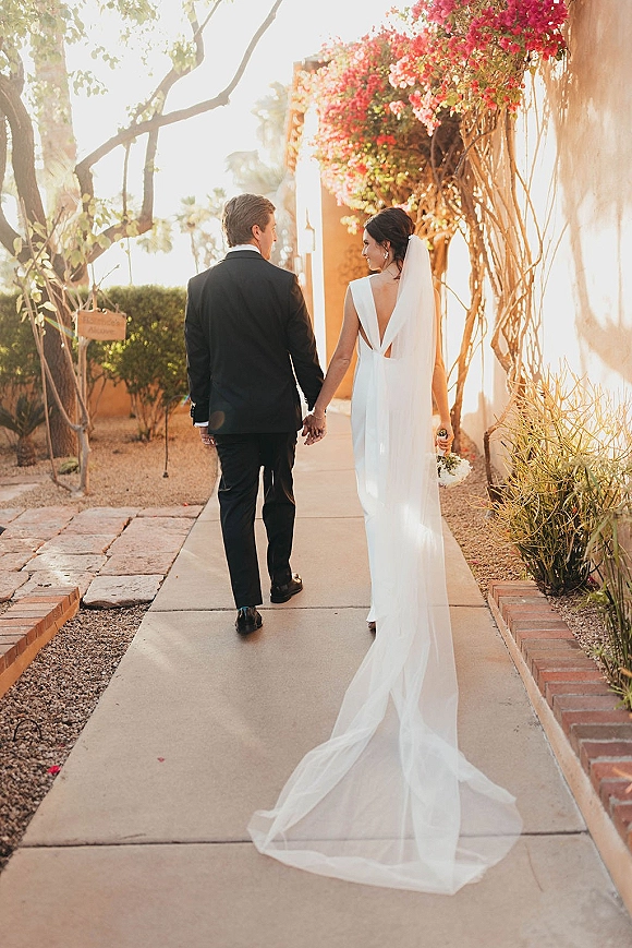 Couple portrait of bride and groom walking hand in hand, bride glancing back with long veil train on a stucco walkway with bougainvillea