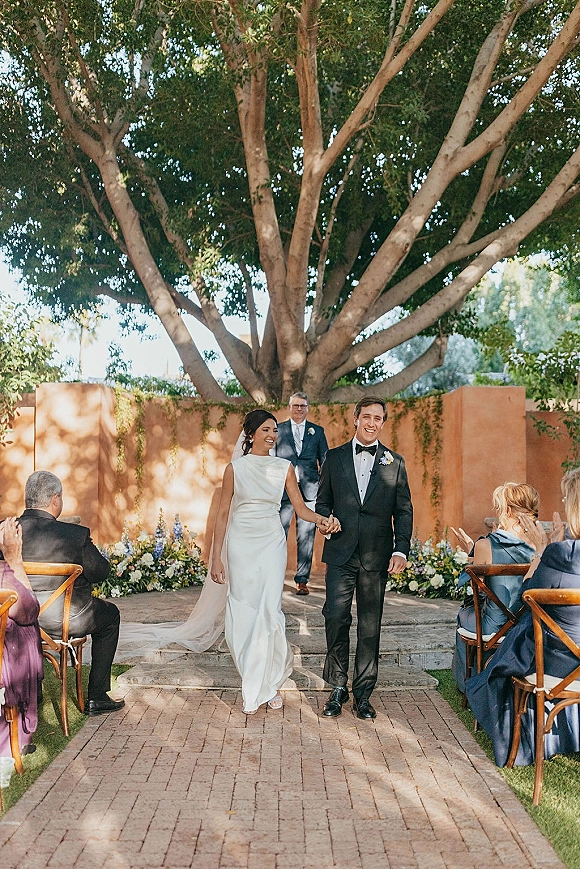 Ceremony recessional as bride and groom walking aisle hand in hand, guests applauding along a brick aisle in an outdoor garden setting